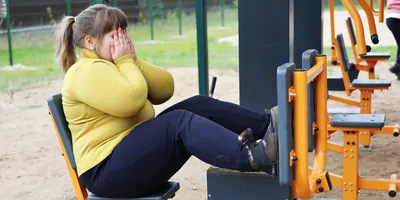 An overweight girl covers her face with hands cupped as she sits on workout equipment in an open area.