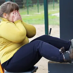 An overweight girl covers her face with hands cupped as she sits on workout equipment in an open area.