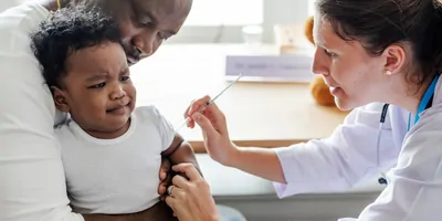 A baby of African descent in an African man's lap gets a first injection.