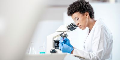A young female scientist wearing PPE looks into a microscope.