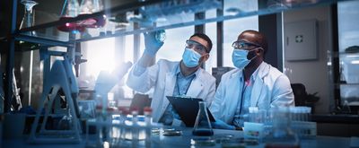 Two men wearing blue masks, goggles, and a white lab coat examine a laboratory sample.
