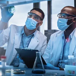 Two men wearing blue masks, goggles, and a white lab coat examine a laboratory sample.