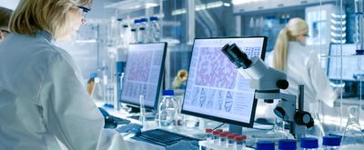 Woman working in laboratory background with blonde hair and white lab coat, microscope and computer with photos of cells featured. 