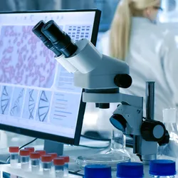 Woman working in laboratory background with blonde hair and white lab coat, microscope and computer with photos of cells featured. 