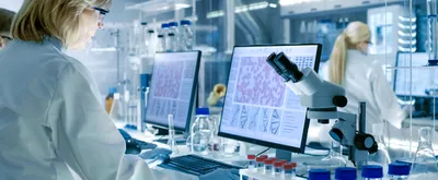 Woman working in laboratory background with blonde hair and white lab coat, microscope and computer with photos of cells featured. 