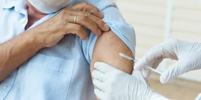 A senior man wearing a protective face mask is getting vaccinated by a clinician wearing white surgical gloves.