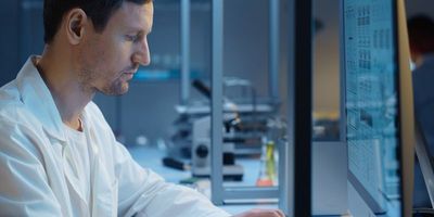 Photograph of lab personnel in a lab coat viewing a desktop in the lab that is displaying some obscured information in a graphical format.