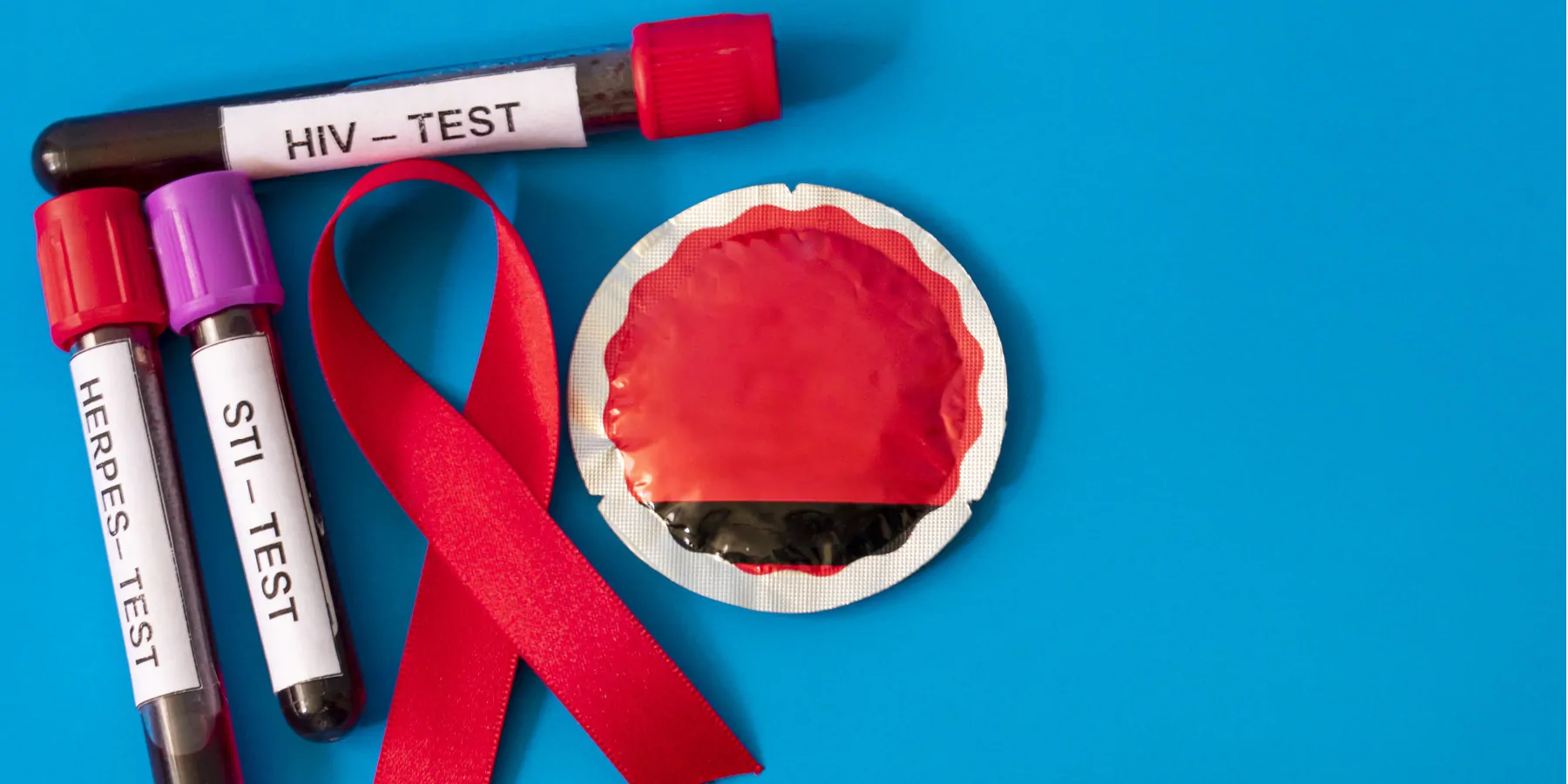 A red ribbon, symbolizing AIDS awareness, with blood tubes from sexually transmitted disease tests and a condom on blue background