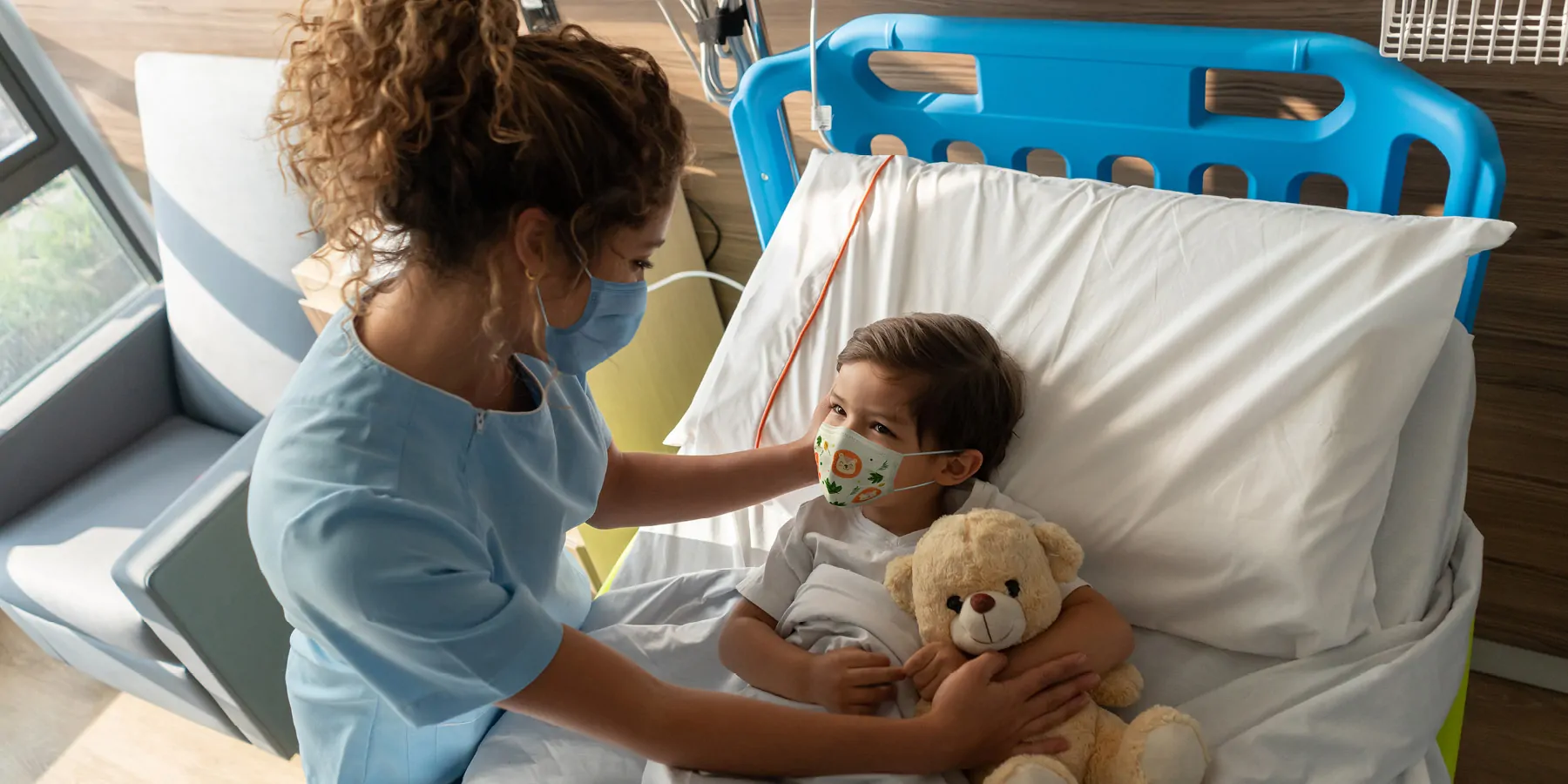 Boy lying down on hospital bed and a nurse comforting him both wearing face masks during the COVID-19 pandemic