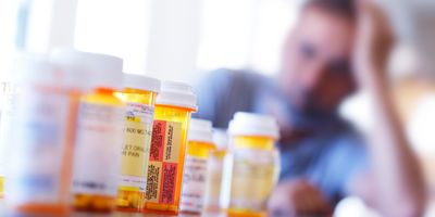 A large group of prescription medication bottles lay on a table in front of a distraught man who is leaning on his hand as he sits at his dining room table.