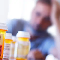 A large group of prescription medication bottles lay on a table in front of a distraught man who is leaning on his hand as he sits at his dining room table.