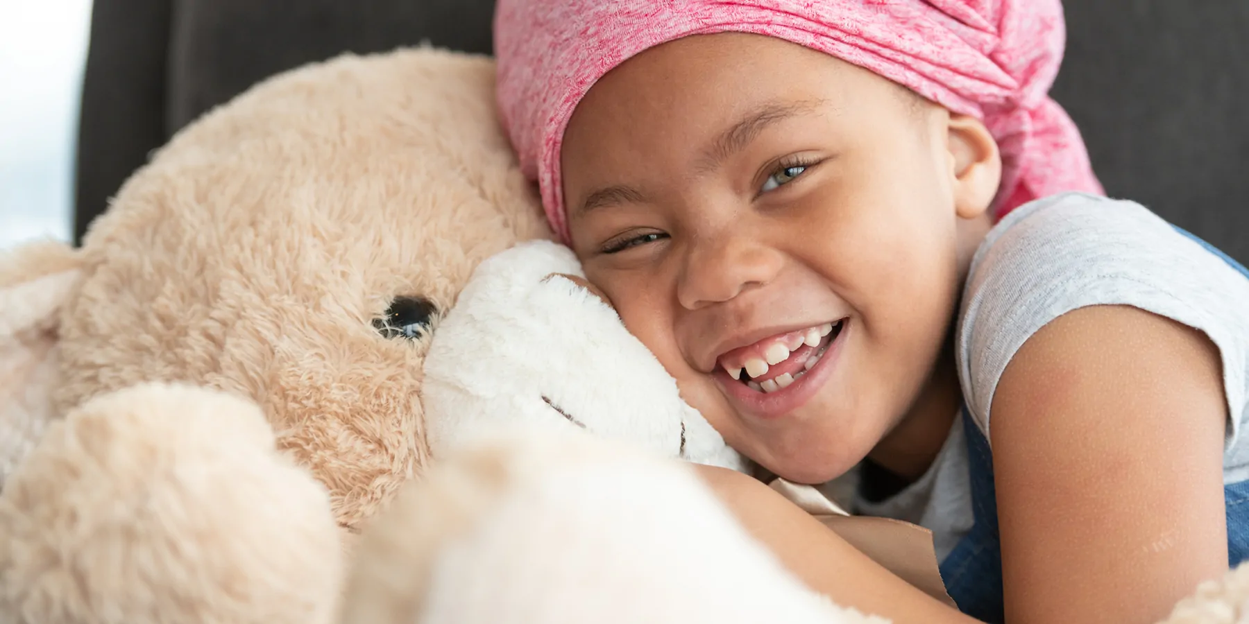 Portrait of a young black girl with cancer. She is wearing a pink bandana and is sitting on a couch. The child is laughing while embracing a large teddy bear.