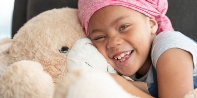 Portrait of a young black girl with cancer. She is wearing a pink bandana and is sitting on a couch. The child is laughing while embracing a large teddy bear.
