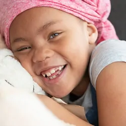 Portrait of a young black girl with cancer. She is wearing a pink bandana and is sitting on a couch. The child is laughing while embracing a large teddy bear.