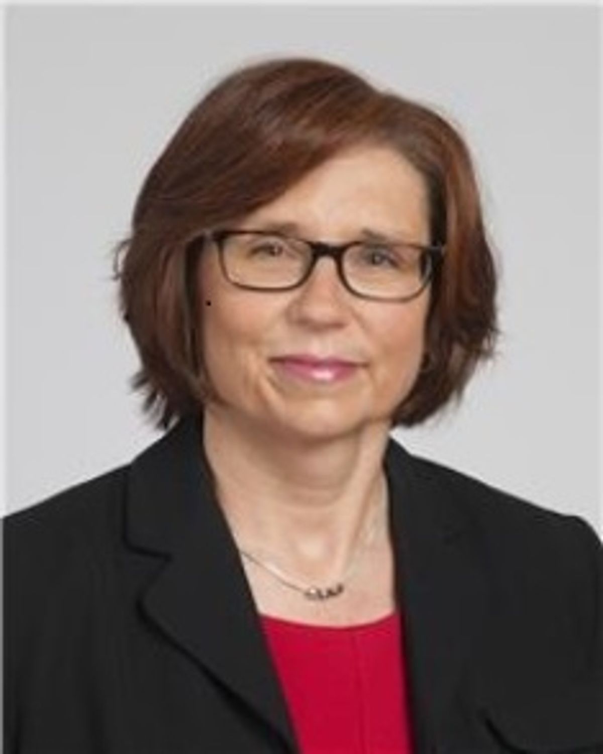 Woman with red hair and black glasses wearing red shirt with black blazer and silver necklace smiles at camera in professional headshot.