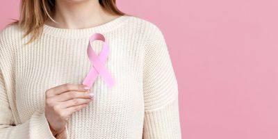 Closeup of woman hand in white pullover holding pink ribbon, the symbol of breast cancer in support of oncology patients.