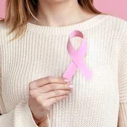 Closeup of woman hand in white pullover holding pink ribbon, the symbol of breast cancer in support of oncology patients.