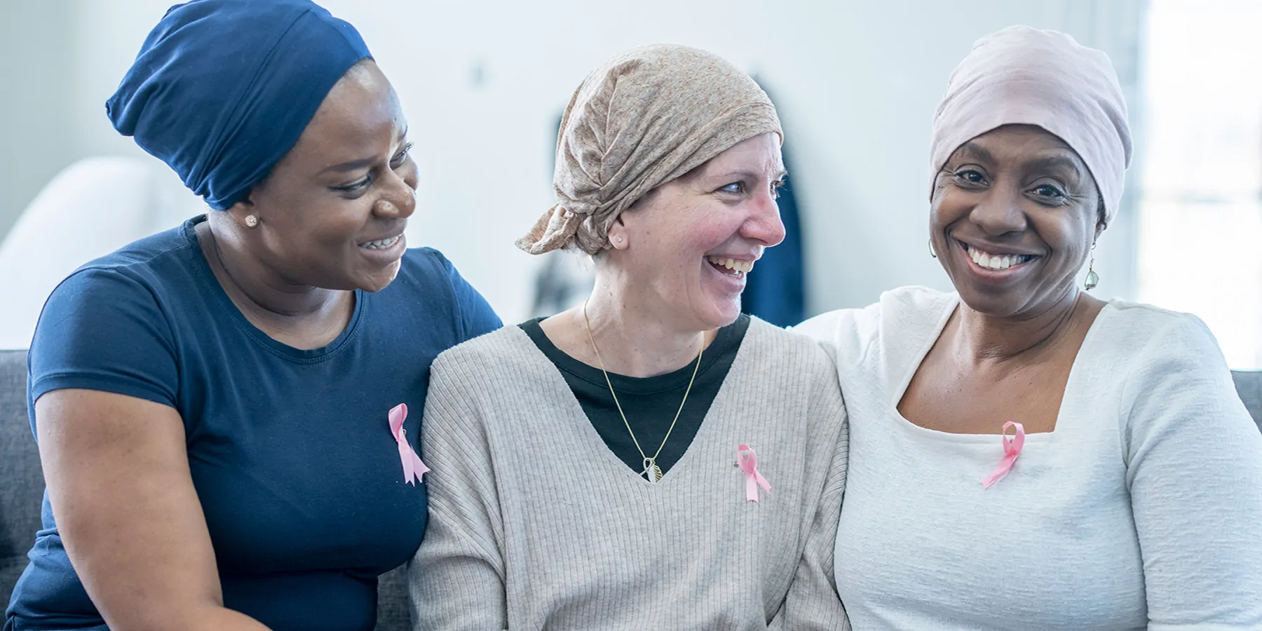 Three women, dressed casually wearing headscarves and pink Breast Cancer awareness ribbons, fighting cancer, sit close to one another on a sofa as they support each other.