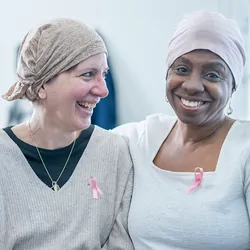 Three women, dressed casually wearing headscarves and pink Breast Cancer awareness ribbons, fighting cancer, sit close to one another on a sofa as they support each other.