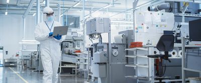 Man in white jumpsuit wearing goggles and black lanyard reads laptop while crossing an engineering lab floor