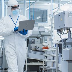 Man in white jumpsuit wearing goggles and black lanyard reads laptop while crossing an engineering lab floor
