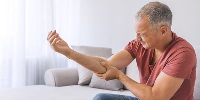 An unhappy mature gray-haired man suffering from muscle pain near his elbow while sitting on a sofa during the day.