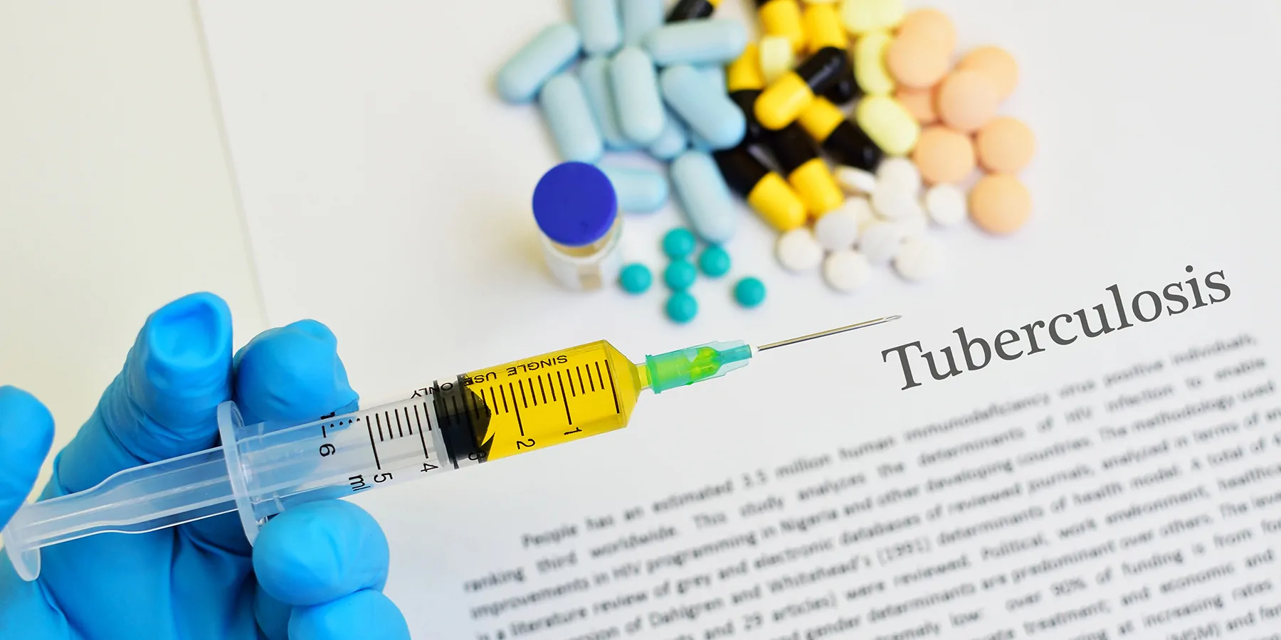 A gloved hand holds a loaded syringe with tablets and capsules and Tuberculosis text printed in the background 