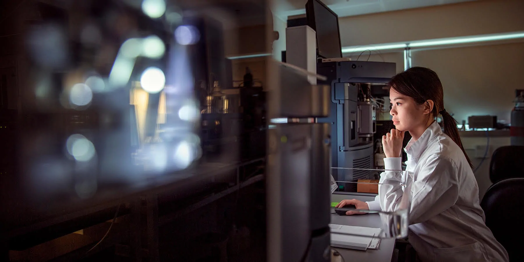 unnamed-1 Dark haired woman with ponytail wearing a white lab coat looks at a computer screen surrounded by black laboratory machines.