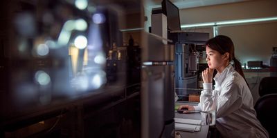 Dark haired woman with ponytail wearing a white lab coat looks at a computer screen surrounded by black laboratory machines.