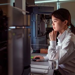 Dark haired woman with ponytail wearing a white lab coat looks at a computer screen surrounded by black laboratory machines.