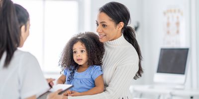 A young mother of African descent, sits in the doctor's office with her toddler on her lap as they discuss the young girl's health