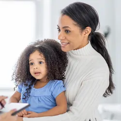 A young mother of African descent, sits in the doctor's office with her toddler on her lap as they discuss the young girl's health