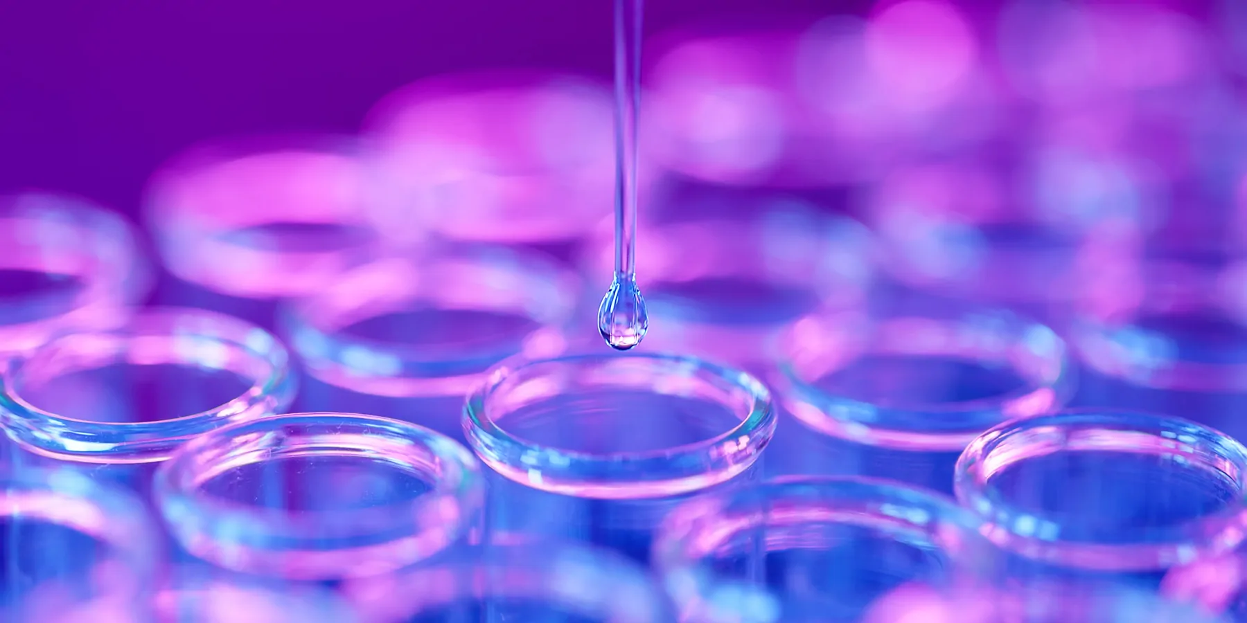 Rows of small glass vials with a pipette tip dripping clear liquid in the center vial against a purple and blue lit background.