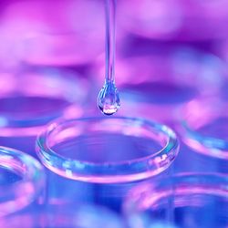 Rows of small glass vials with a pipette tip dripping clear liquid in the center vial against a purple and blue lit background.
