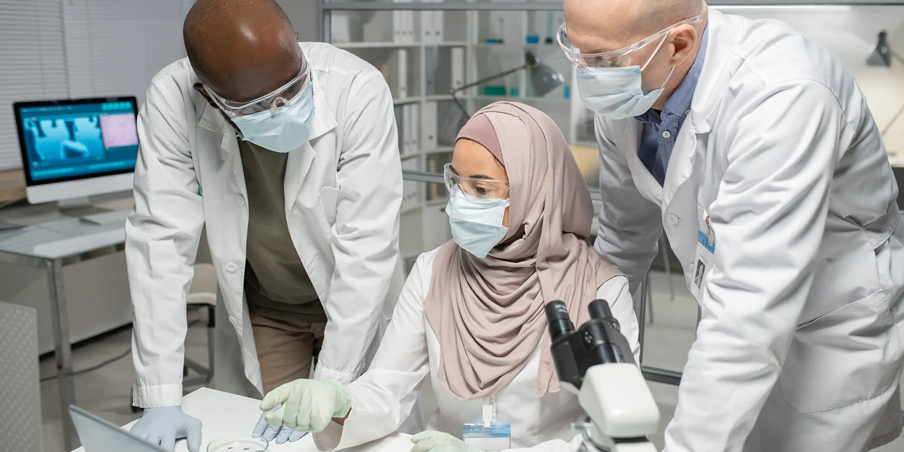 Two interracial male scientists in PPE listening to a female colleague wearing a hijab and PPE.