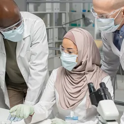 Two interracial male scientists in PPE listening to a female colleague wearing a hijab and PPE.