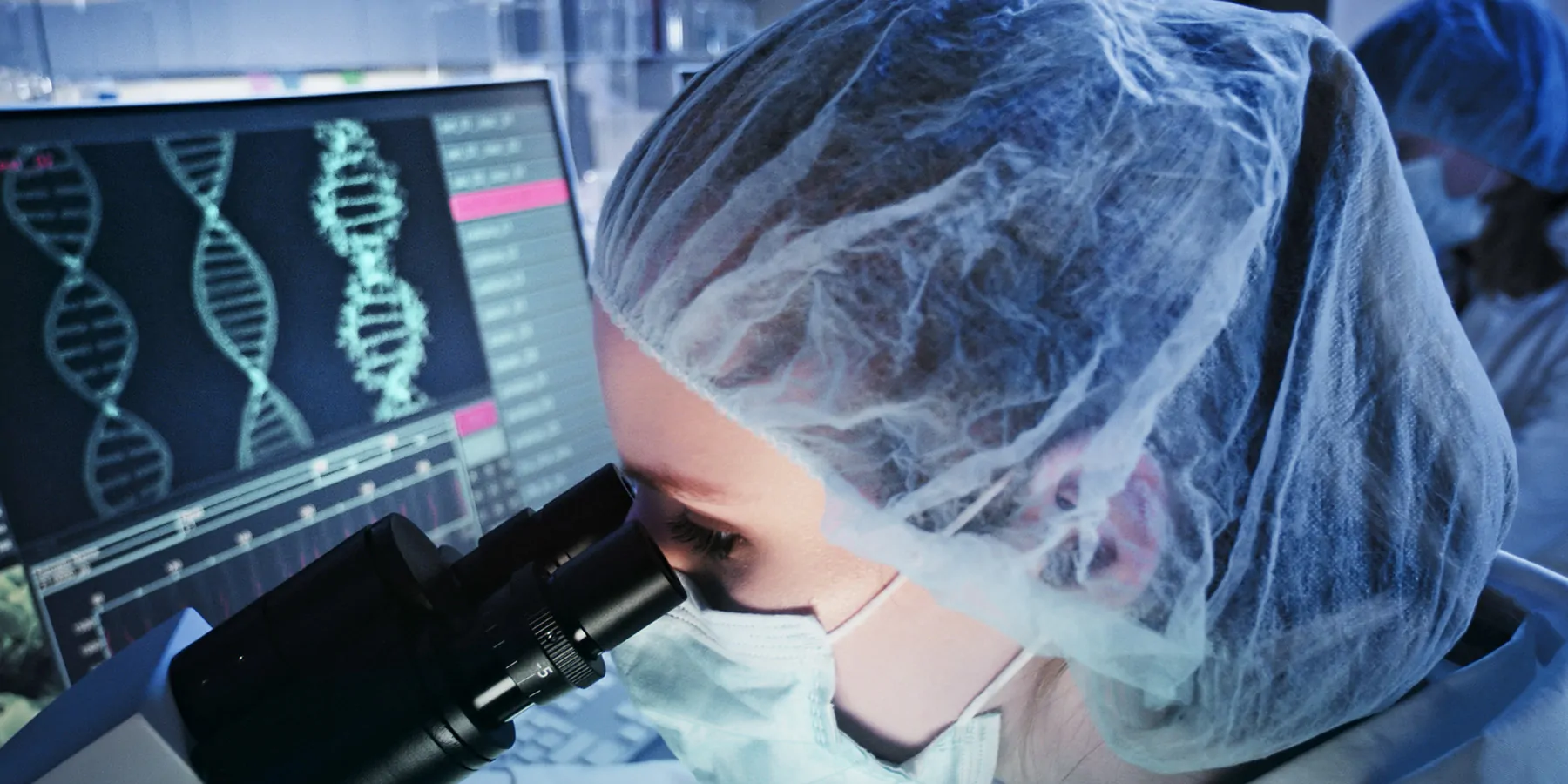 A female researcher studying a specimen under a microscope with computer screens showing DNA helix in the background