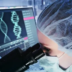 A female researcher studying a specimen under a microscope with computer screens showing DNA helix in the background