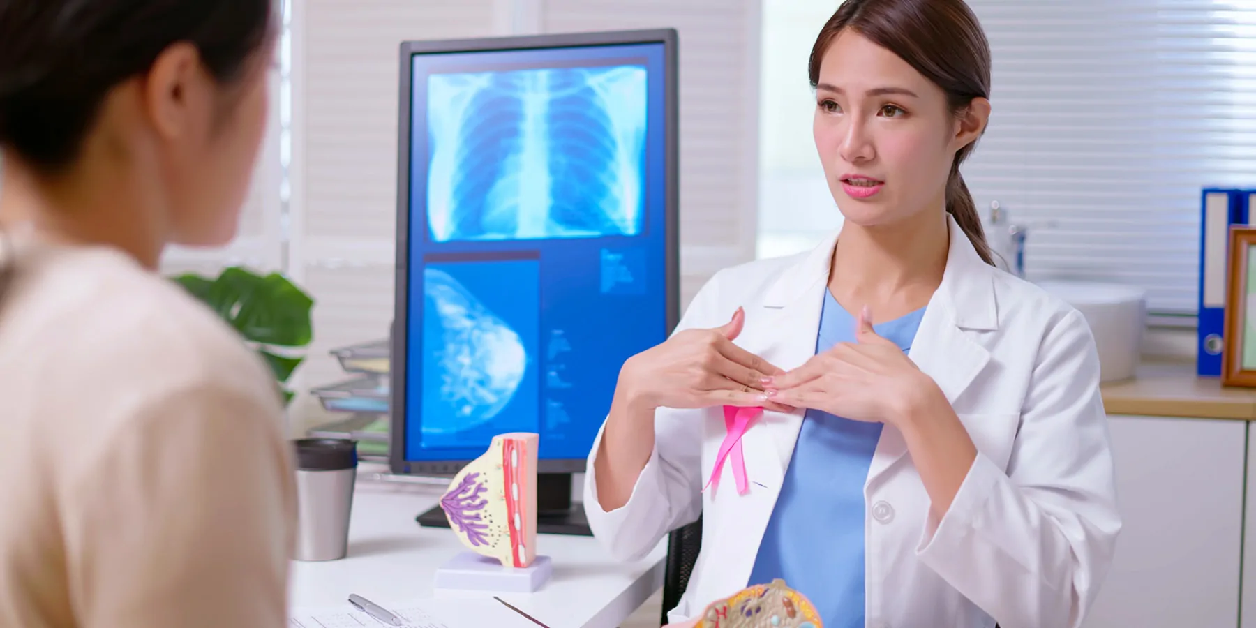 A female doctor wearing a white coat with a pink ribbon explaining breast self-examination to a female patient at a clinic.