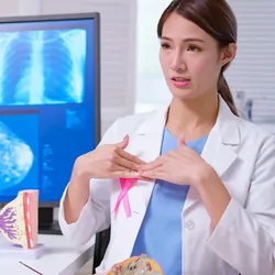 A female doctor wearing a white coat with a pink ribbon explaining breast self-examination to a female patient at a clinic.