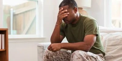 An adult male soldier covers his eyes with his hand as he sits alone on a couch.