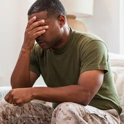 An adult male soldier covers his eyes with his hand as he sits alone on a couch.