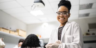 Young woman with dark skin and black curly hair in a bun is wearing a white lab coat, white gloves, and black rimmed goggles smiling at the camera in a lab.