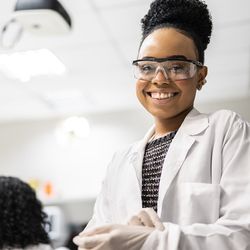 Young woman with dark skin and black curly hair in a bun is wearing a white lab coat, white gloves, and black rimmed goggles smiling at the camera in a lab. 
