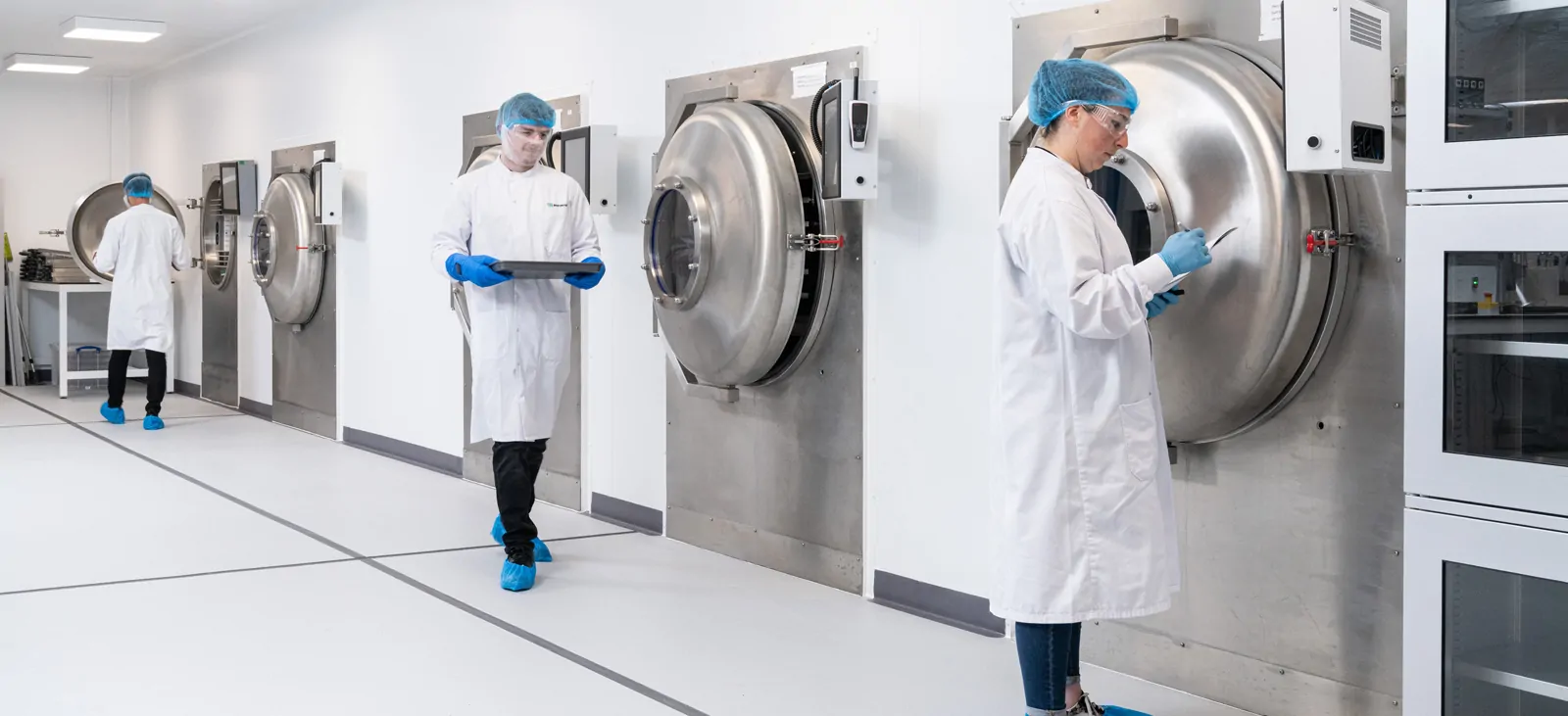 Women in white lab coat, blue hair net, and blue gloves stands in front of industrial laboratory freezer/lyophilizer. 