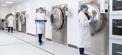Women in white lab coat, blue hair net, and blue gloves stands in front of industrial laboratory freezer/lyophilizer.