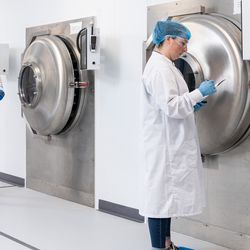 Women in white lab coat, blue hair net, and blue gloves stands in front of industrial laboratory freezer/lyophilizer. 