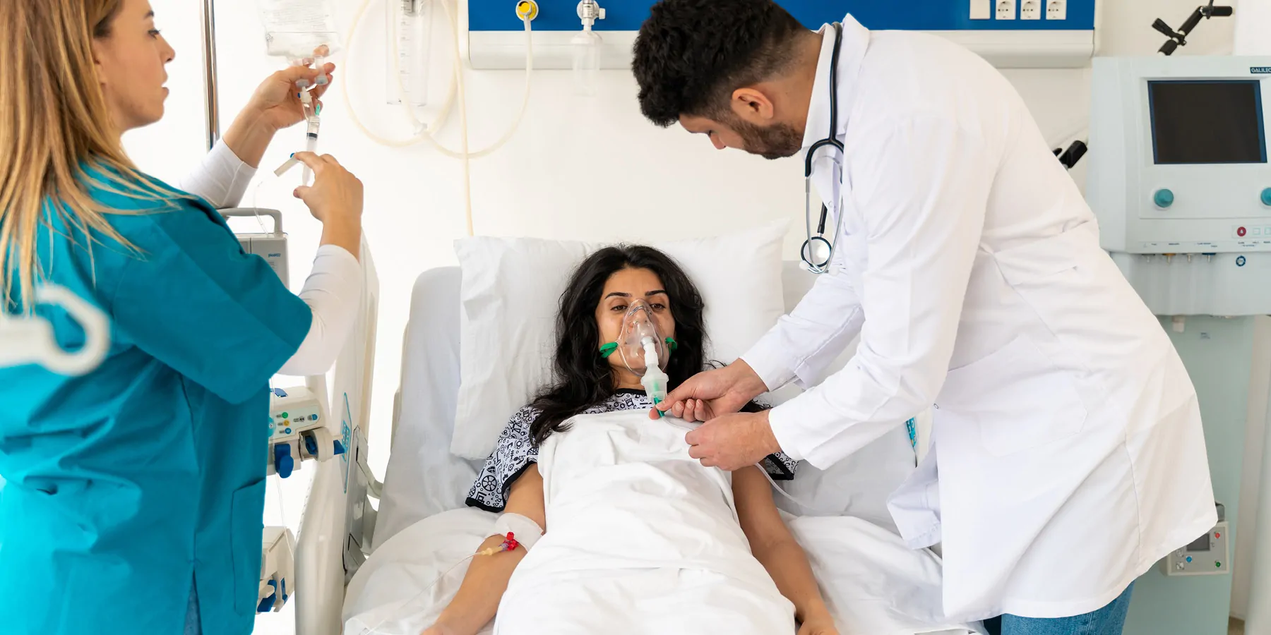 Clinicians with protective masks examine the infected female patient in the ICU who rests on a bed with an oxygen mask.