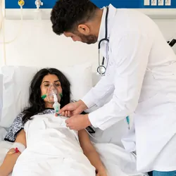 Clinicians with protective masks examine the infected female patient in the ICU who rests on a bed with an oxygen mask.