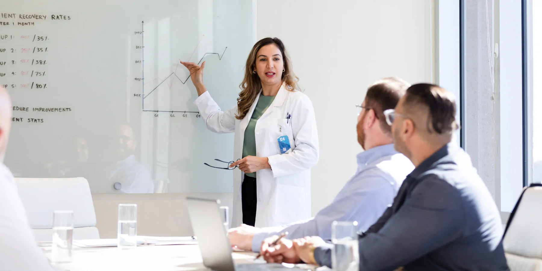 A female clinician points to a graph on the board as she explains the data represented to the seated male members.